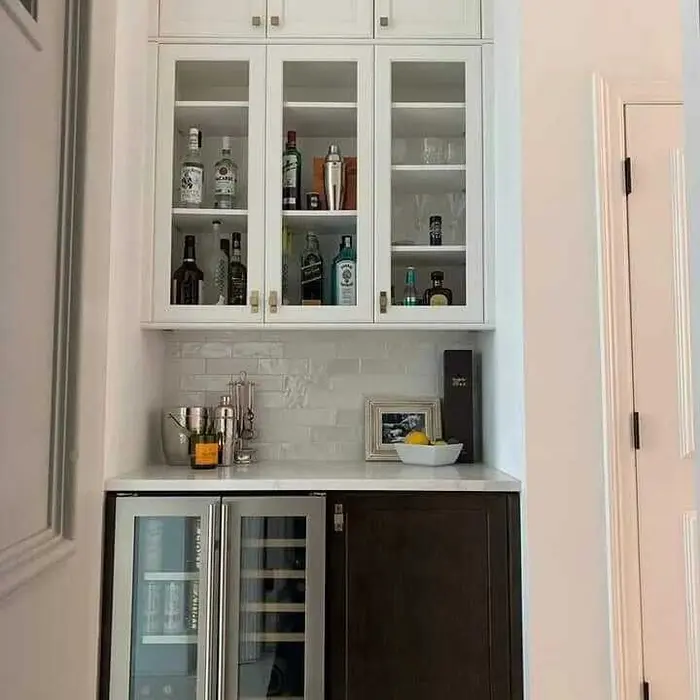 Barra de cocina moderna completada con gabinetes superiores de vidrio en blanco que exhiben bebidas alcohólicas, mosaico gris en la salpicadura, encimera de cuarzo blanco, y refrigerador de vino/bebidas dual debajo