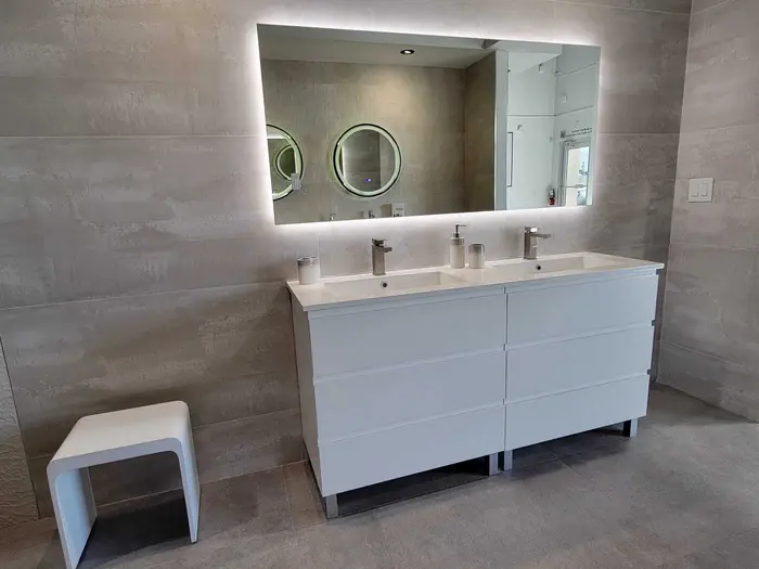 Modern bathroom with double white vanity, LED-backlit mirror, concrete-look tile walls, and matching stool
