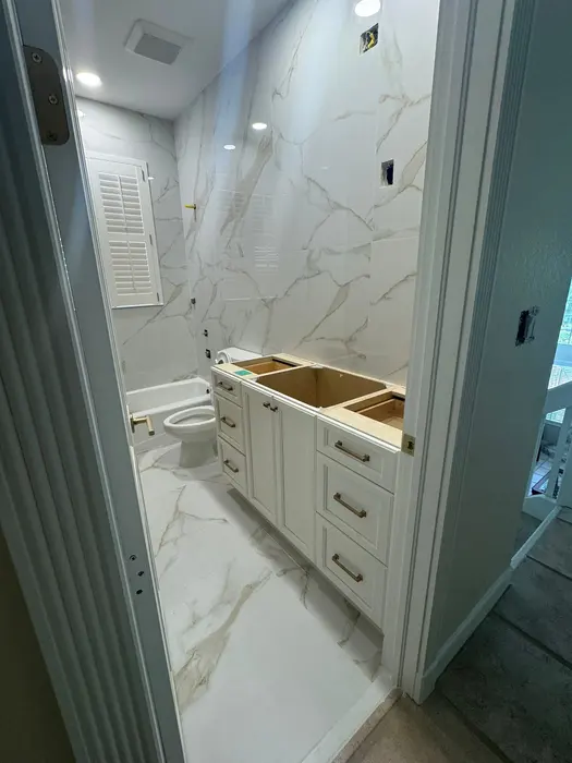 A bathroom during renovation showing marble-look tile walls and floor, white cabinetry base without countertop, recessed lighting, and toilet installation in progress.