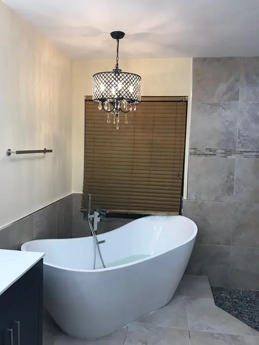 Completed modern bathroom with freestanding white oval soaking tub, gray tile walls, bronze pendant light fixture with crystal details, and sliding wooden-slat window shade