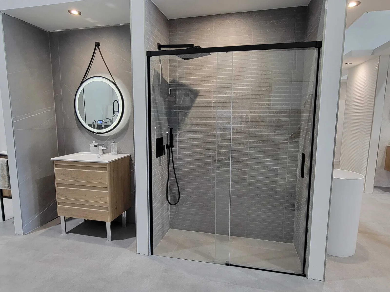 Modern bathroom with light wood vanity, round black-framed mirror, gray subway tile walls, and walk-in shower with glass enclosure