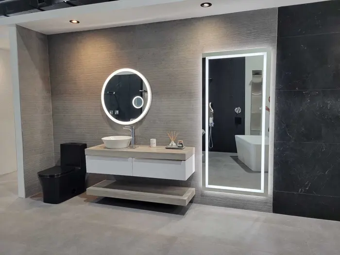 Modern bathroom showroom display featuring a white vessel sink vanity with LED mirror, black toilet, freestanding white bathtub, textured gray tile walls, and black accent tiles