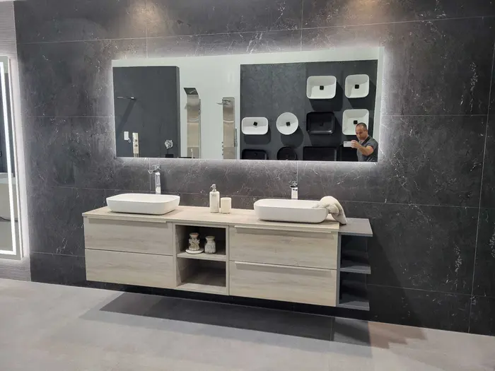 Bathroom vanity display showroom with double vessel sinks, natural wood cabinetry, dark tile backdrop, and wall-mounted fixture samples