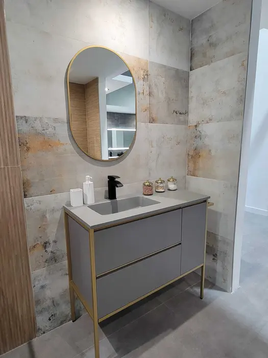 Modern bathroom vanity with gray cabinetry, brass hardware, white countertop sink, and oval brass-framed mirror against textured tile walls