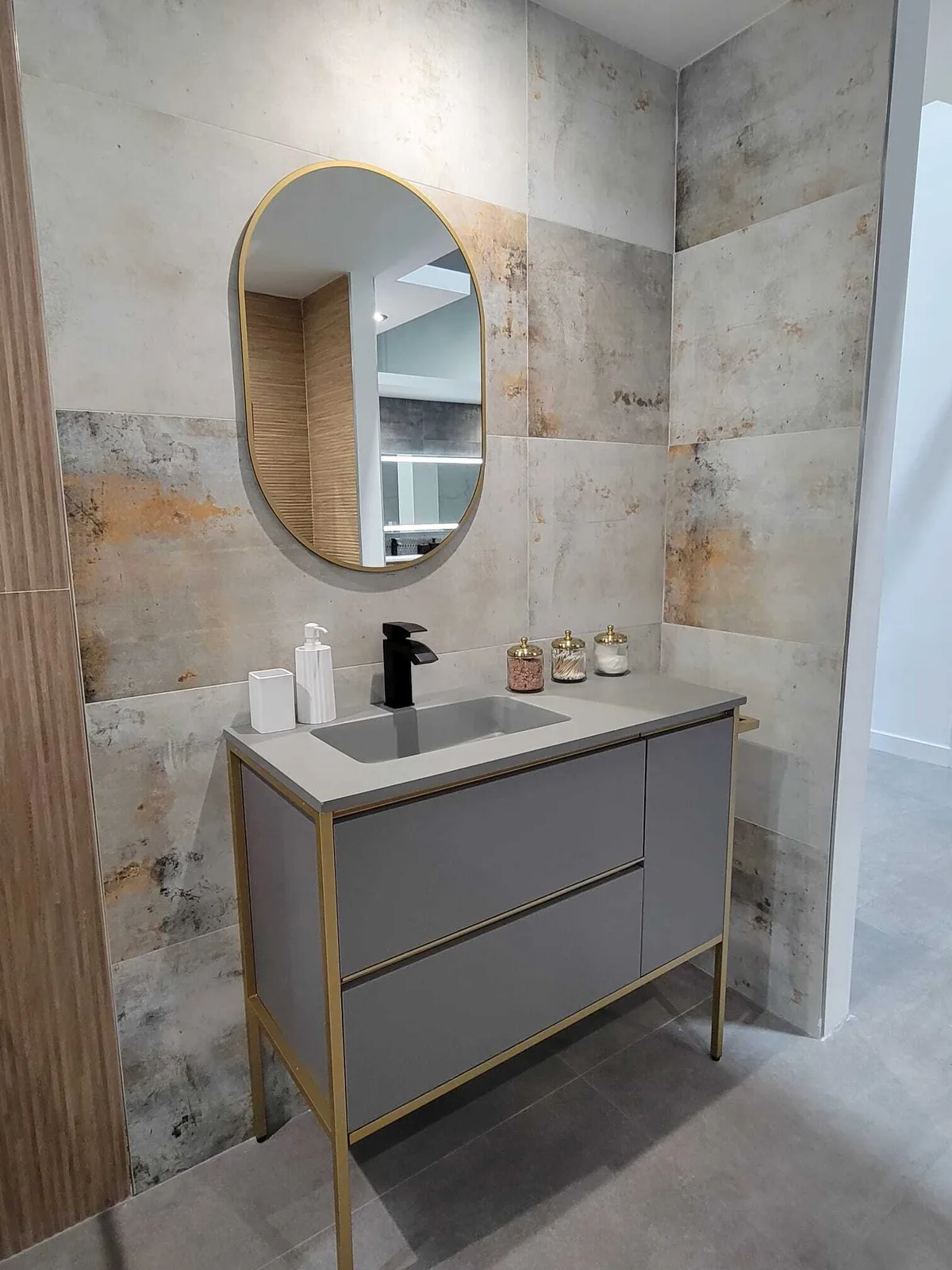 Modern bathroom vanity with gray cabinetry, brass hardware, white countertop sink, and oval brass-framed mirror against textured tile walls