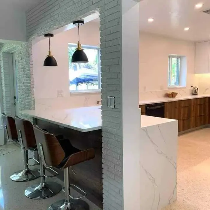 Completed modern kitchen featuring a white marble-topped island with seating, green brick accent wall, pendant lighting, and wood cabinetry