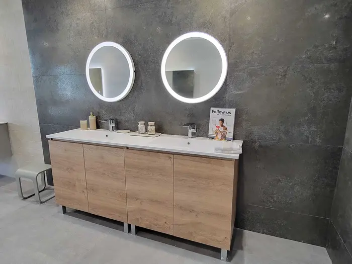 Showroom display of a modern bathroom vanity with dual sinks, white quartz countertop, natural wood cabinetry, and contemporary LED-backlit round mirrors against a dark tile accent wall.