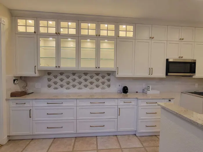 Modern white kitchen with freshly painted cabinetry, glass-front upper cabinets, decorative fan-pattern backsplash tile, and light countertops in a residential home
